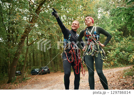 Male is showing something by hand and talking. Man and woman doing climbing in forest with use of safety equipment Male is showing something by hand and talking. Man and woman doing climbing in forest with use of safety equipment 129786814