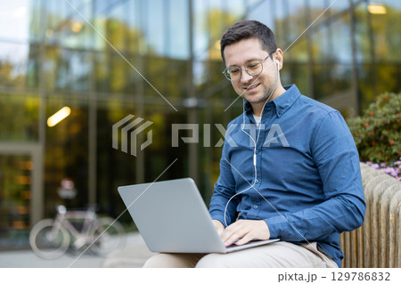 A professional adult man sitting outdoors, wearing earphones, working on a laptop, surrounded by greenery and modern architecture, exuding productivity and engagement in a comfortable and natural A professional adult man sitting outdoors, wearing earphones, working on a laptop, surrounded by greenery and modern architecture, exuding productivity and engagement in a comfortable and natural 129786832