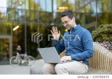 A young professional man engaging in a video call using a laptop, sitting outdoors on a wooden bench in front of a modern glass building with nearby flowers and bicycle. A young professional man engaging in a video call using a laptop, sitting outdoors on a wooden bench in front of a modern glass building with nearby flowers and bicycle. 129786833