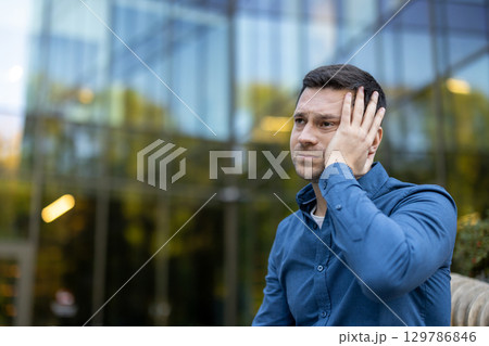 A man dressed in a blue shirt looks worried while standing outside with a modern glass facade blurred in the background. The scene evokes thoughtfulness, reflection, and a sense of concern. 129786846