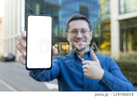 Smiling man outdoors holding a blank smartphone screen towards the camera while giving a thumbs up, indicating satisfaction or showcasing space for customizable content. Smiling man outdoors holding a blank smartphone screen towards the camera while giving a thumbs up, indicating satisfaction or showcasing space for customizable content. 129786859