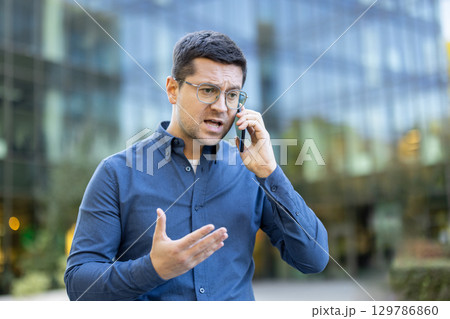 A concerned male in glasses talks on a smartphone, gesturing emphatically, outdoors near a glass office building. A concerned male in glasses talks on a smartphone, gesturing emphatically, outdoors near a glass office building. 129786860