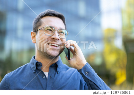 A cheerful professional man engaged in a phone conversation near a glass building exterior A cheerful professional man engaged in a phone conversation near a glass building exterior 129786861