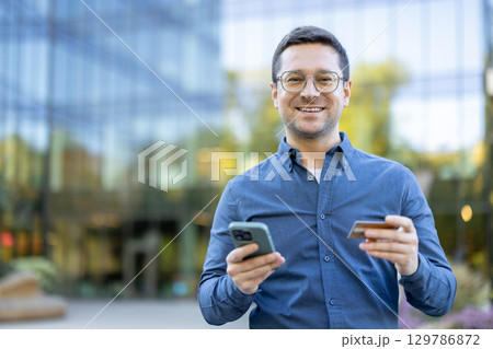 Cheerful man holding a smartphone and credit card while outdoors in a business environment. Cheerful man holding a smartphone and credit card while outdoors in a business environment. 129786872