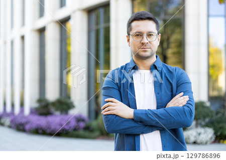 A portrait of a confident young man standing with arms crossed outdoors. Modern architecture and landscaped surroundings provide a contemporary atmosphere to the scene, reflecting professionalism 129786896