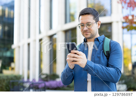A man wearing glasses and a backpack is using his smartphone outdoors near modern architecture. A man wearing glasses and a backpack is using his smartphone outdoors near modern architecture. 129786897