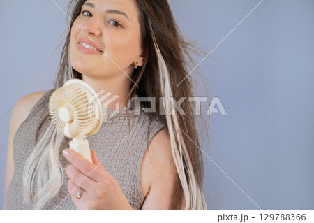 Young woman holding a compact fan feeling refreshed and cool outdoors on a hot sunny summer day. 129788366