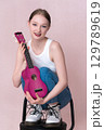 Playful woman musician holding ukulele and squatting with her feet up on bar stool. Positive woman with smile on face looking at camera, playfully posing with stringed musical instrument, having fun 129789619