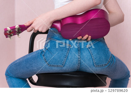 Rear view of sitting female musician holding ukulele behind her back. Sexy unrecognizable woman in blue tight jeans that hugs her figure. Back view of midsection female body holding musical instrument 129789620