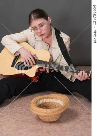 Sad woman guitarist street musician playing acoustic guitar and looking hopefully at camera. Young woman sits with legs apart and empty straw hat lies in front of her for donation for playing guitar 129789654