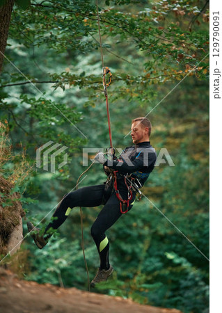 Up on the rope, hanging. Man is doing climbing in the forest by use of safety equipment Up on the rope, hanging. Man is doing climbing in the forest by use of safety equipment 129790091
