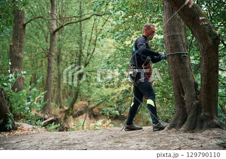 Standing on the ground and preparing. Man is doing climbing in the forest by use of safety equipment Standing on the ground and preparing. Man is doing climbing in the forest by use of safety equipment 129790110