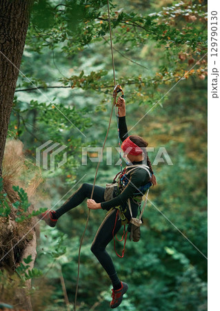 In sportive clothes, high up on the rope. Woman is doing climbing in the forest by the use of safety equipment 129790130