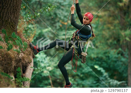 Looking to the side, hanging on the rope. Woman is doing climbing in the forest by the use of safety equipment 129790137