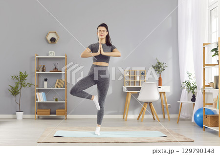 Relaxed slim woman practicing yoga in Vrksasana tree pose at home, meditating with hands in namaste. 129790148