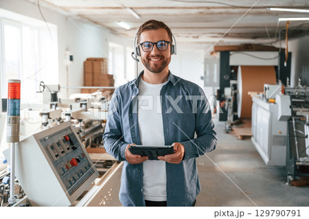 In headphones, with tablet. Handsome man is working at the factory of creating eco boxes In headphones, with tablet. Handsome man is working at the factory of creating eco boxes 129790791