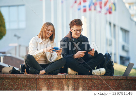 Smiling, having fun. Two young students are sitting outdoors against university Smiling, having fun. Two young students are sitting outdoors against university 129791149