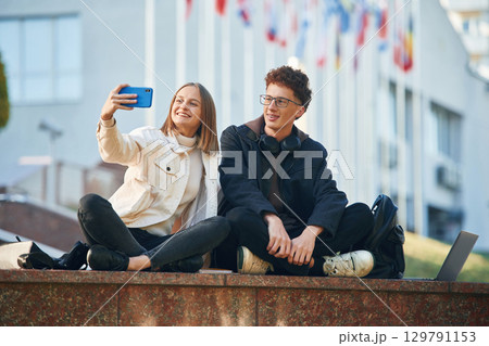 Holding smartphone. Two young students are sitting outdoors against university Holding smartphone. Two young students are sitting outdoors against university 129791153