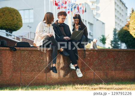 Sitting and using laptop. Three young students are outside the university outdoors Sitting and using laptop. Three young students are outside the university outdoors 129791226