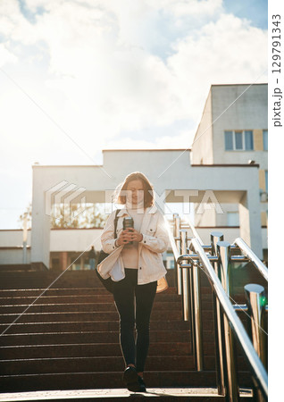 Illuminated by sunlight, holding bottle of water. Young female student in casual clothes is outdoors 129791343