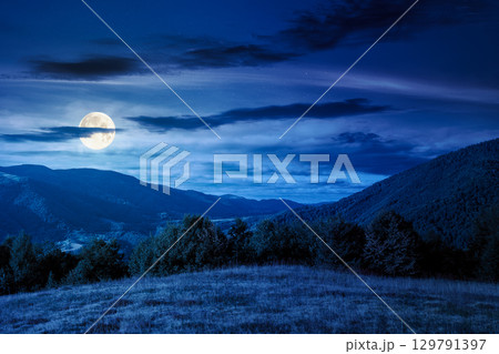 mountain landscape with forest on the hill in autumn at night. beautiful view in to the rural valley in full moon light. alpine countryside scenery under sky with clouds mountain landscape with forest on the hill in autumn at night. beautiful view in to the rural valley in full moon light. alpine countryside scenery under sky with clouds 129791397