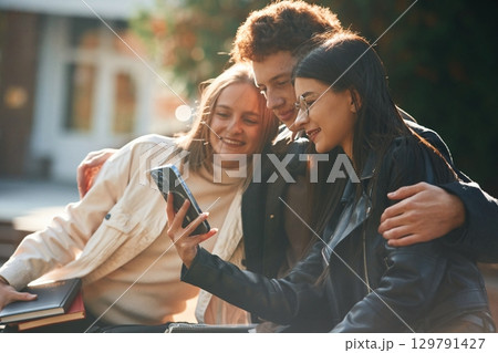 Girl is showing something in smartphone. Three young students are outside the university outdoors 129791427