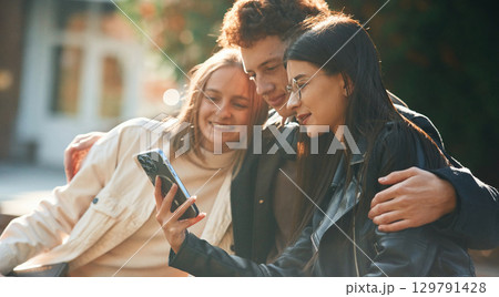 Girl is showing something in smartphone. Three young students are outside the university outdoors Girl is showing something in smartphone. Three young students are outside the university outdoors 129791428