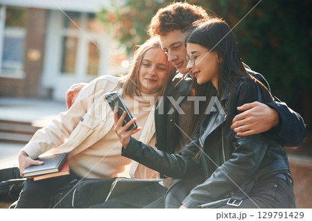 Girl is showing something in smartphone. Three young students are outside the university outdoors 129791429
