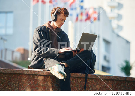 In headphones, using laptop. Young student is outside the university, in black clothes 129791490