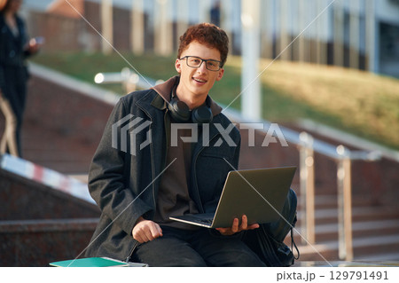 Smiling, holding laptop. Young student is outside the university, in black clothes 129791491