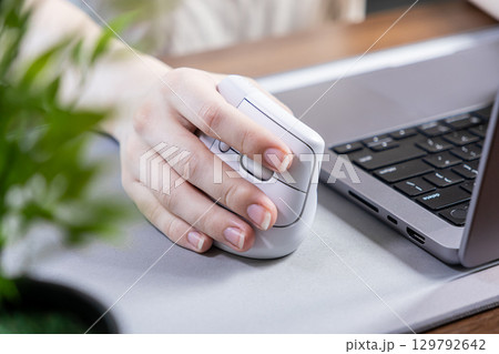 A person's hand comfortably gripping an ergonomic vertical mouse next to a laptop, promoting wrist health and efficient digital work in a modern office setup A person's hand comfortably gripping an ergonomic vertical mouse next to a laptop, promoting wrist health and efficient digital work in a modern office setup 129792642