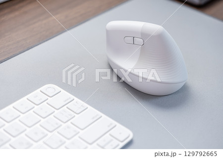 Modern white ergonomic vertical computer mouse sits on a grey desk mat next to a keyboard, emphasizing comfortable workspace setup and digital user health 129792665