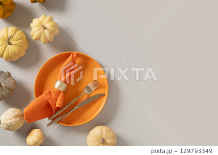 Autumn and Thanksgiving Day table setting with white small pumpkin, orange empty plate with golden cutlery on gray background. Concept catering. Top view. Copy space. Autumn and Thanksgiving Day table setting with white small pumpkin, orange empty plate with golden cutlery on gray background. Concept catering. Top view. Copy space. 129793349