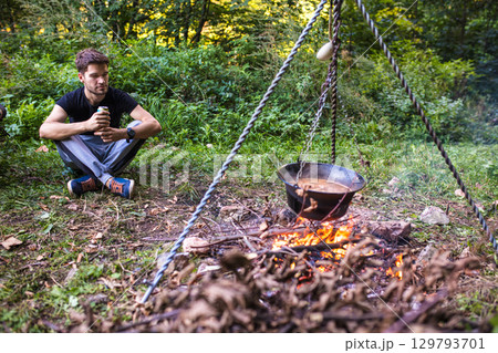 Hiker relaxing while cooking on campfire in forest 129793701