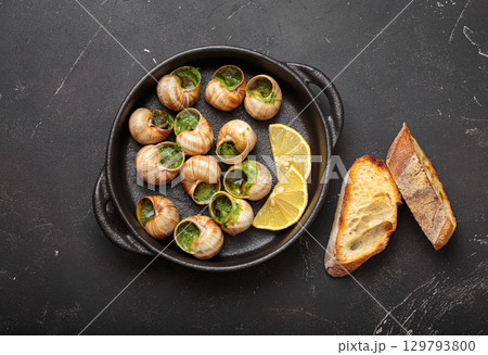 Snails escargots with garlic butter and lemon, served in a black pan with toasted bread baguette slices on black rustic background Snails escargots with garlic butter and lemon, served in a black pan with toasted bread baguette slices on black rustic background 129793800