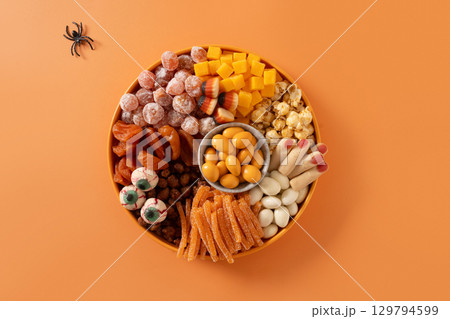 Halloween sweet charcuterie board with pumpkin, popcorn, dried fruits, color chocolate candy, spider, crackers on orange background. Festive food for kids party. Top view. 129794599