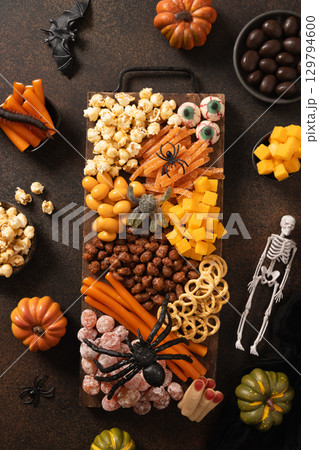 Halloween sweet charcuterie board with pumpkin, popcorn, color chocolate candy, spider, crackers with scary decoration on dark background. Festive food for kids party. View from above. Halloween sweet charcuterie board with pumpkin, popcorn, color chocolate candy, spider, crackers with scary decoration on dark background. Festive food for kids party. View from above. 129794600