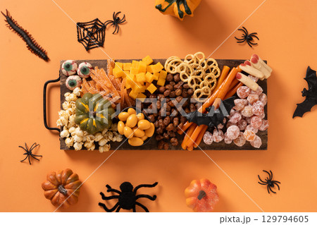 Halloween sweet charcuterie board with pumpkin, popcorn, dried fruits, color chocolate candy, spider, crackers on orange background. Festive food for kids party. View from above. 129794605