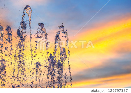 Water fountain splashes against a vibrant sunset sky with colorful clouds Water fountain splashes against a vibrant sunset sky with colorful clouds 129797587