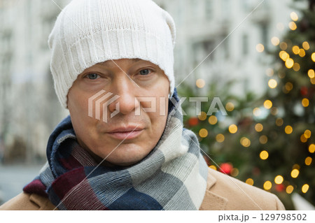 Portrait of man wearing white beanie and cozy scarf, with blurred Christmas festive background. Cold winter weather. Portrait of man wearing white beanie and cozy scarf, with blurred Christmas festive background. Cold winter weather. 129798502