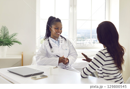 Friendly smiling woman doctor talking with female patient sitting at the desk in medical office. 129800545