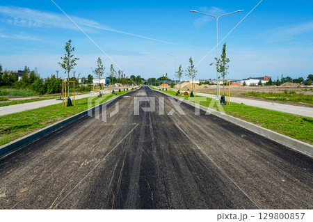 Newly paved asphalt road with sidewalks, young trees, and street lighting in modern urban development 129800857
