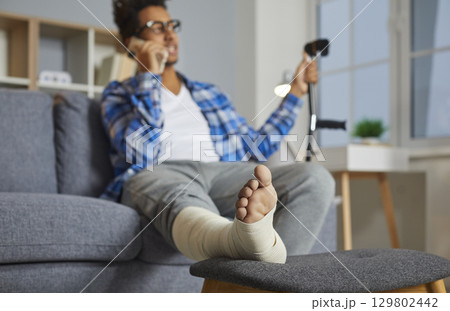 Man talking on phone while sitting on couch, with his injured foot resting on stool 129802442