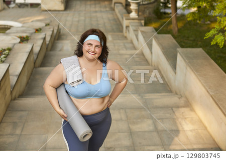 Happy chubby woman in sportswear standing on park steps, holding workout mat and smiling Happy chubby woman in sportswear standing on park steps, holding workout mat and smiling 129803745