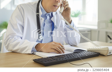 Medical professional doctor, male nurse at office desk, talking on phone, consulting notebook 129805404
