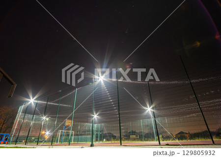 Outdoors mini football and basketball court with ball gate and basket surrounded with high protective fence brightly illuminated with spotlight lamps at night. 129805932
