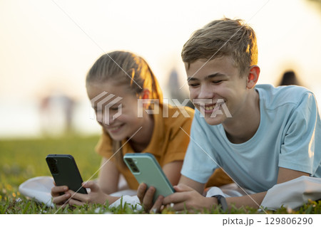 Teens texting with their friends on cellphones outdoors in summer park. Teens texting with their friends on cellphones outdoors in summer park. 129806290