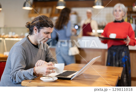 Middle aged man sitting at table, using laptop and enjoing cup of tea in coffee house. Modern lifestyle concept Middle aged man sitting at table, using laptop and enjoing cup of tea in coffee house. Modern lifestyle concept 129811018
