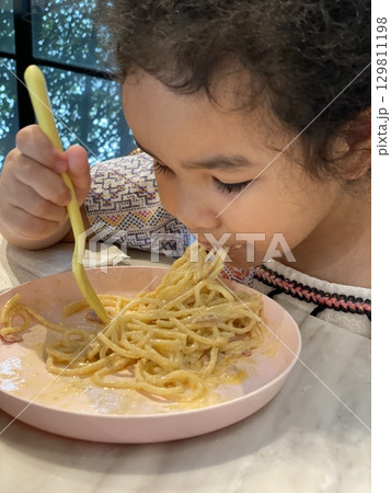 Close up view of little girl eating delicious spaghetti carbonara with a fork in a restaurant. 129811198