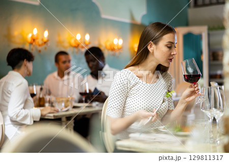 Positive young woman eating salad and drinking red wine in cozy restaurant Positive young woman eating salad and drinking red wine in cozy restaurant 129811317